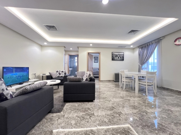 Modern living room with grey sofas, a wall-mounted TV, and a white dining table near a window with gray curtains.