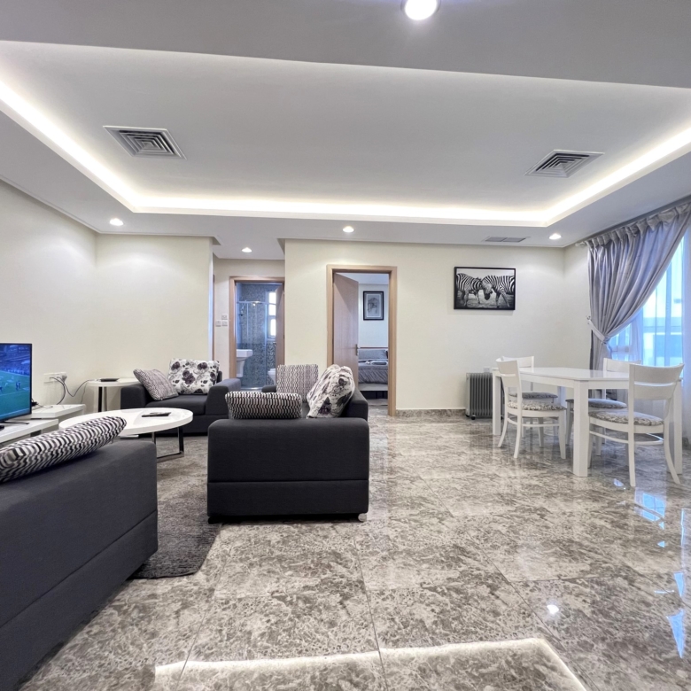 Modern living room with grey sofas, a wall-mounted TV, and a white dining table near a window with gray curtains.